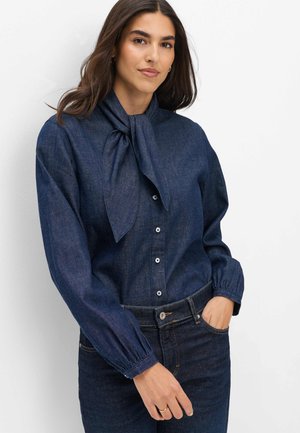 Kvinde iført en mørkeblå denimbluse med bundet sløjfe ved halsen og matchende denimjeans, stående med den ene hånd i lommen.