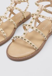 Sandalias planas beige con tachuelas doradas decorativas en las correas, que cuentan con una plantilla de color marrón suave y una suela marrón, mostrando un diseño de correas texturizadas.