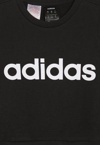 Czarny sweterek z okrągłym dekoltem, z dużym białym logo "adidas" na piersi. Gładka struktura materiału. Metka pielęgnacyjna widoczna wewnątrz.