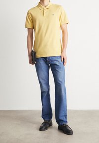 Gul kortärmad pikétröja, blå jeans och svarta loafers. Pikén har en krage och en liten logodetalj på bröstet.