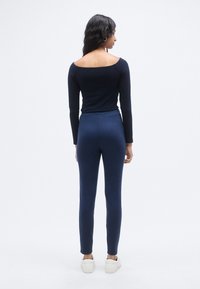 Μπλε σκούρο μπλουζάκι με μακριά μανίκια και σχεδιασμό έξω από τον ώμο, σε συνδυασμό με εφαρμοστά μπλε σκούρα παντελόνια και λευκά αθλητικά παπούτσια. Λείο υλικό και κομψή σιλουέτα.