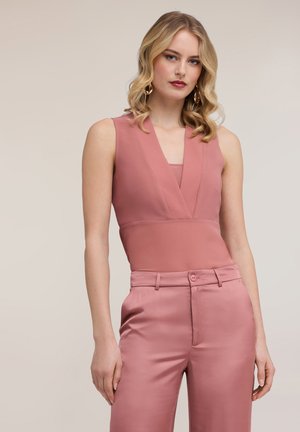 Blond žena v bezrukávovém topu s výstřihem do V v barvě zaprášené růže a ladicích saténových kalhotách s vysokým pasem, pózující na neutrálním pozadí.