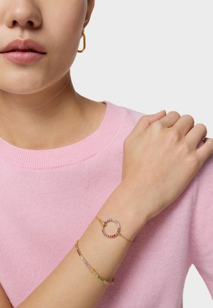 Mujer con un suéter rosa y dos pulseras doradas, una circular con piedras multicolores y otra delgada con piedras multicolores.
