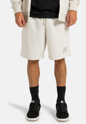Beige katoenen shorts met een elastische tailleband, zijzakken en een subtiele logo op de onderste zoom. G gedragen met zwarte sokken en schoenen.