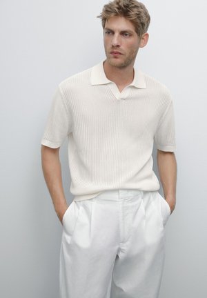 Beige ribbad stickad polo med en liten krage, matchad med vita veckade byxor. Materialen verkar mjuka och avslappnade i passformen.
