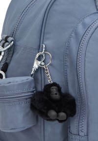 Grå stof rygsæk med lynlås lomme, prydet med en sort gorilla-formet nøglepude lavet af blødt materiale og plastdetaljer.