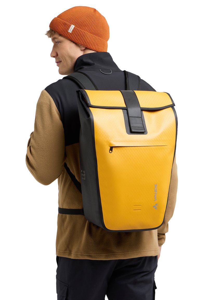 Sac à dos étanche jaune et noir avec une texture géométrique, fermeture à enroulement et poche zippée à l'avant. Sangles ajustables et détail du logo visibles.