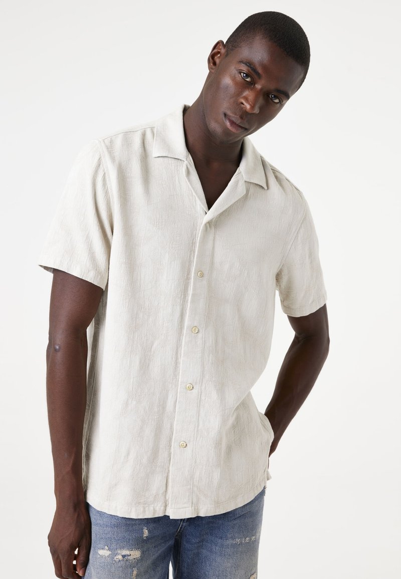 Korte mouwen, lichtbeige shirt met een gestructureerd patroon en een camp-kraag. Voorzien van witte knopen en een relaxte pasvorm. Gepaard met vervaagde blauwe spijkerbroeken.