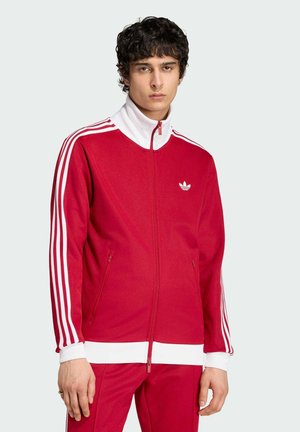 Młody mężczyzna w czerwonej dresowej kurtce Adidas i pasujących spodniach z białymi paskami, stojący na jednolitym jasnym tle.