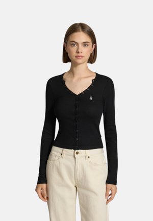 Giovane donna che indossa un top nero aderente a manica lunga con bottoni e scollo a V e pantaloni beige chiaro a vita alta su uno sfondo semplice.