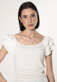 Witte gebreide top met gefronde mouwen, een scalloped halslijn en horizontale perforaties langs het lichaam voor textuur en detail.