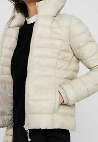 Veste matelassée beige clair avec capuche, poignets élastiques et fermeture éclair frontale. Tissu texturé avec motif matelassé horizontal.