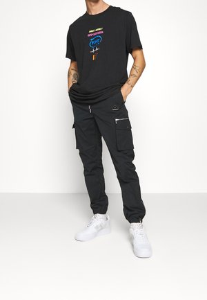 T-shirt noir avec un graphique coloré, associé à un pantalon cargo noir doté de fermetures éclair et de poches, complété par des baskets blanches.