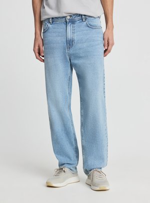 Osoba ubrana w jasnoniebieskie jeansy o prostych nogawkach i jasnoszare trampki, stojąca na tle jednolitego białego tła.