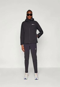 Person stands wearing black hooded jacket, matching black pants, white sneakers, and large black sunglasses against plain background.