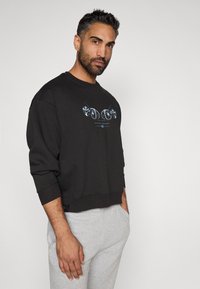 Sweatshirt preto com design cropped, punhos canelados e decote redondo. A estampa gráfica na frente apresenta elementos circulares em azul e branco.