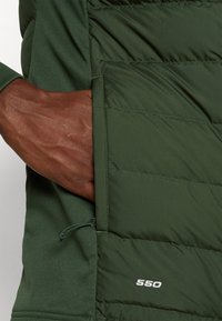 Main à la peau foncée insérée dans la poche zippée d'une veste isolante vert foncé avec une couture "550" sur le panneau avant.