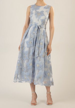 Ärmelloses hellblaues Midikleid mit floralem Spitzenmuster, metallischen Akzenten, gebundenem Taillengürtel und silbernen offenen Absatzschuhen am Modell.