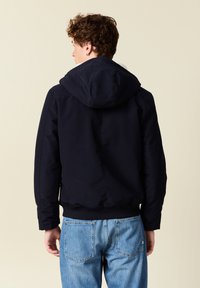 Giubbotto bomber blu navy con cappuccio, chiusura a zip e polsini a costine, abbinato a jeans azzurri, visto da dietro.