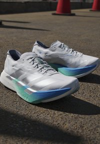 Een paar witte hardloopschoenen met blauw-groene verloopzolen geplaatst op een buitenverharding in het zonlicht.