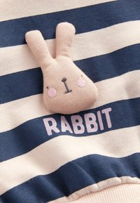 Visage de lapin en peluche beige doux avec des joues roses sur tissu rayé bleu marine et crème, avec le mot « RABBIT » imprimé en dessous.
