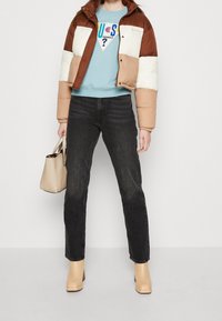 Kvinna iklädd färgblockerad brun och beige pufferjacka, ljusblå sweatshirt med logotyp, svarta jeans, beige stövlar, hållande beige handväska.