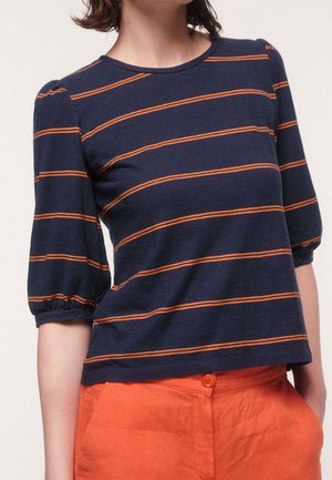 Kobieta ubrana w granatową koszulę z poziomymi pomarańczowymi paskami i bufkami o długości do łokcia, połączoną z pomarańczowymi spodniami.