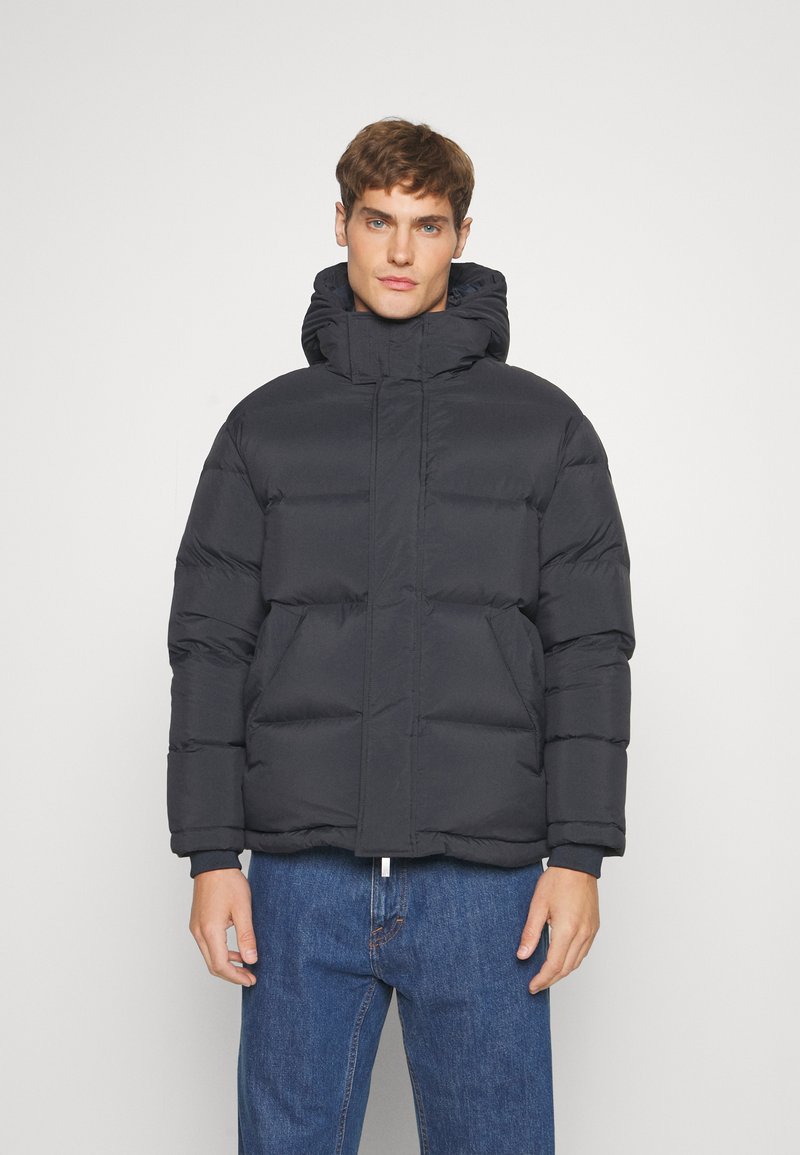 Black puffer jacket with a high collar and hood, featuring a quilted design, visible stitching, and a front zipper. Worn with blue jeans.