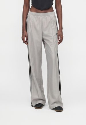 Personne à la peau noire portant un pantalon de survêtement large gris clair avec des bandes noires et un haut court noir, debout sur fond blanc.