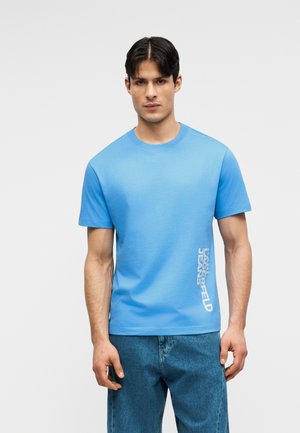 Muž v svetlomodrom tričku s krátkym rukávom s bielym vertikálnym nápisom "Karl Lagerfeld Jeans" na boku, doplnený o modré džínsy.