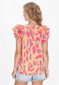 Blusa a maniche corte in tessuto arancione con motivi astratti rosa e blu; dettagli arricciati sulle spalle; abbinata a pantaloni corti di denim.