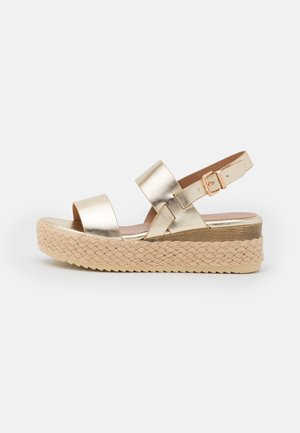 Sandale compensée avec brides métalliques dorées, boucle réglable et semelle plateforme en jute tressée sur fond blanc.