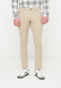 Pantalon en coton beige à coupe slim, avec une fermeture à boutons, deux poches avant et des détails de couture discrets. Assorti à des baskets blanches et grises.