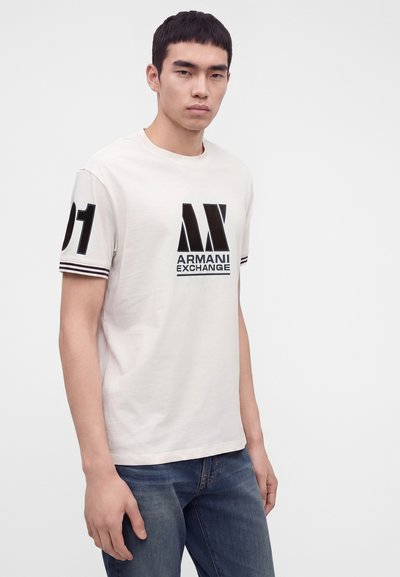 Млад мъж с къса тъмна коса, облечен в бяла тениска Armani Exchange с черно лого и номер 01 на ръкава, комбинирана със сини дънки.