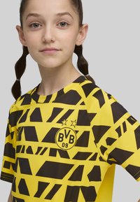 Camiseta juvenil con un patrón geométrico amarillo y negro, mangas cortas, cuello redondo y un logo con "BVB 09" y dos estrellas.