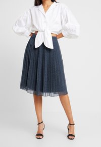 Camicia bianca a maniche lunghe con bottoni annodata in vita, abbinata a una gonna midi plissettata blu navy con piccoli pois bianchi. Tacchi neri con cinturini.