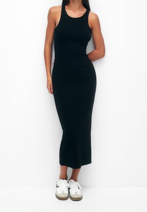 Femme en robe midi noire moulante sans manches et baskets blanches, debout devant un fond blanc.