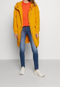 Parka impermeable amarilla con cordones, dos bolsillos frontales y un corte relajado, combinada con una camiseta texturada naranja y jeans azules con acentos verdes.