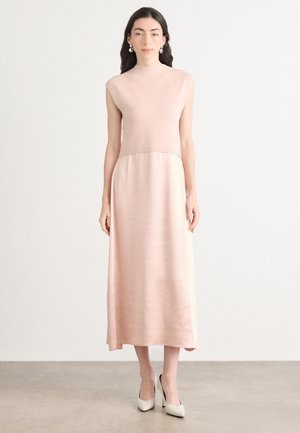 Robe midi sans manches rose avec un haut ajusté, une jupe fluide et une texture lisse. Assortie à des talons blancs et des boucles d'oreilles simples.