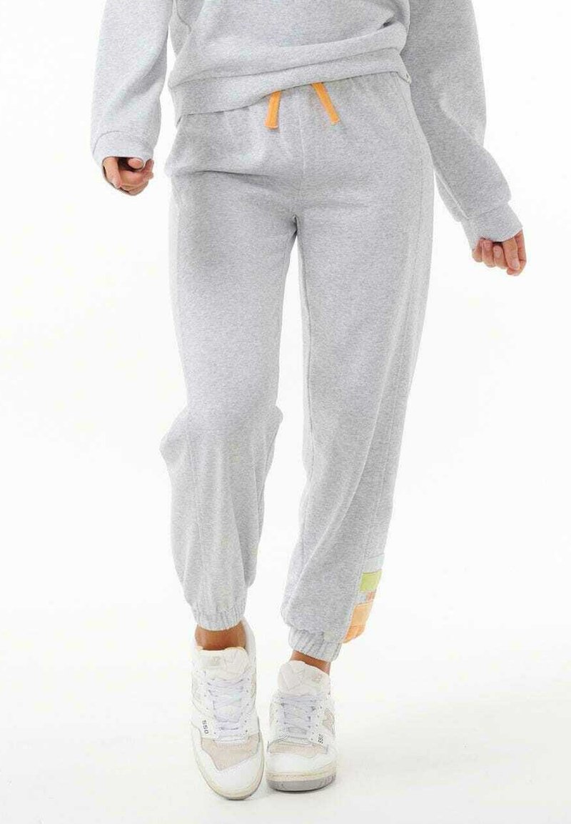 Persona con pantalones jogger gris claro con puños elásticos y cordón naranja, combinados con zapatillas blancas, de pie contra un fondo blanco.