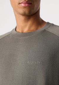 Gros plan sur une personne portant un sweat-shirt olive avec col rond, arborant un logo Marc O'Polo subtilement brodé sur la poitrine.