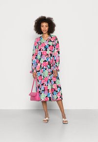 Vestido midi floral com mangas longas, apresentando padrões vibrantes de rosas em rosa, verde e azul sobre um fundo preto. Acessório: bolsa rosa.