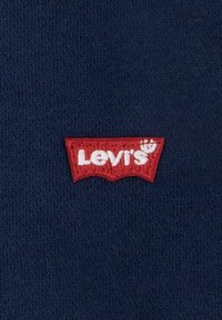 Tissu bleu marine avec un patch logo Levi's rouge brodé en forme de sweat à capuche stylisé. Le texte est blanc, avec un bord inférieur incurvé.