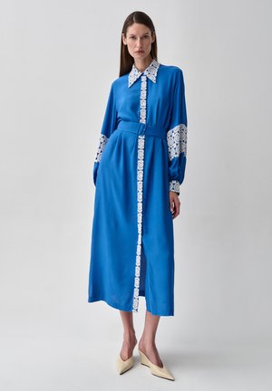 Femme portant une robe midi bleue avec ceinture et bordure en dentelle blanche sur le col, le devant et les manches, se tenant devant un fond clair uni.