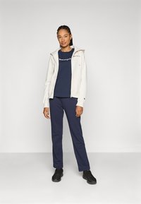 Svijetlosmeđa zip-up majica s kapuljačom i vezicama, tamnoplava majica s bijelim logotipom, tamnoplave trenirke i crne sportske cipele. Glatka tekstura tkanine.
