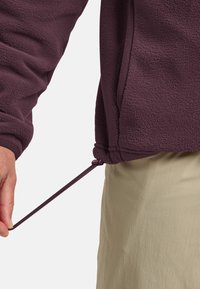 Fleece pullover in dark purple with a textured surface; features a drawstring at the hem held by a hand against beige fabric below.