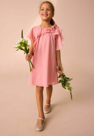Rosa jurk met ruches aan de mouwen, tot op de knie. Kind houdt groene bloemen vast, draagt zilveren ballerina's. Zachte stof en vloeiende ontwerp.