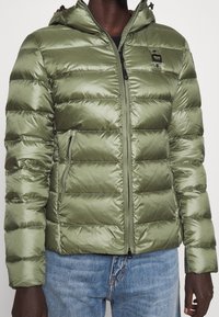 Zielona kurtka puchowa z kapturem, wykonana z błyszczącego, pikowanego materiału. Posiada kieszenie zapinane na zamek oraz zamek błyskawiczny z przodu. Gruba, ocieplona konstrukcja.