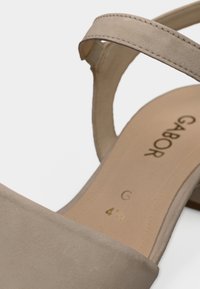 Μπεζ σανδάλι με απαλό επάνω μέρος που μοιάζει με σουέντ, διαθέτει ένα λουράκι και χαμηλό τακούνι. Χρυσή σήμανση ορατή στην εσωτερική σόλα. Απλός σχεδιασμός.