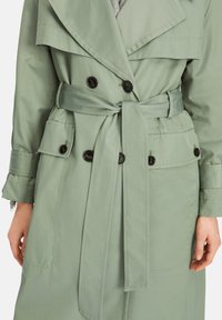 Light green trench coat with a double-breasted front, large lapels, a belted waist, and two buttoned pockets. Smooth, water-resistant fabric.
